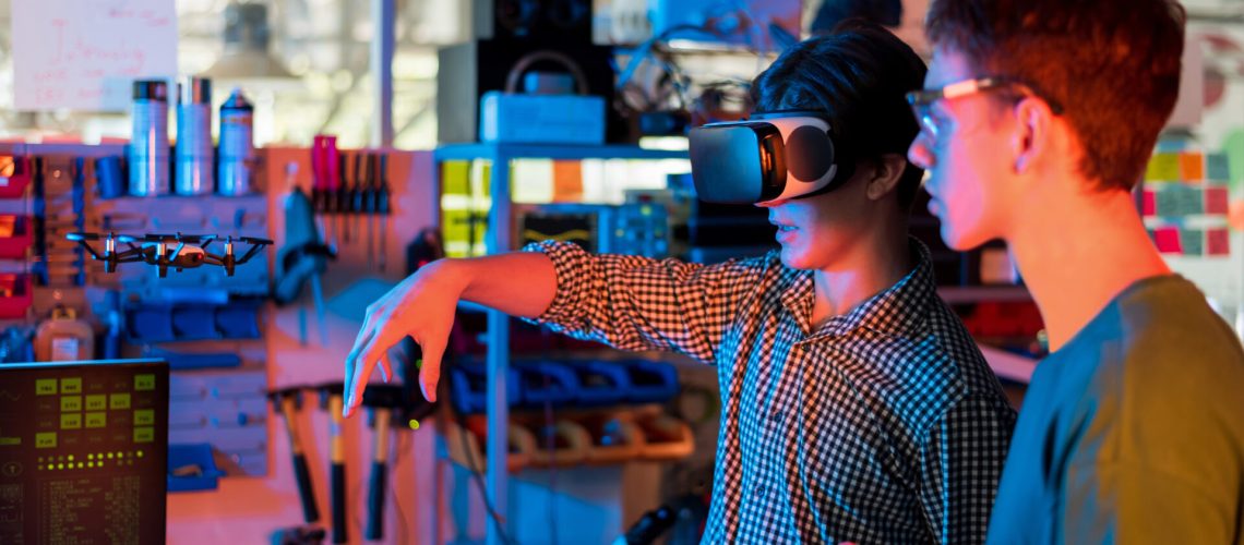Teens doing experiments in robotics in a laboratory. Boy in protective glasses and girl in VR headset controlling a flying drone using controller and hand. Red and blue illumination